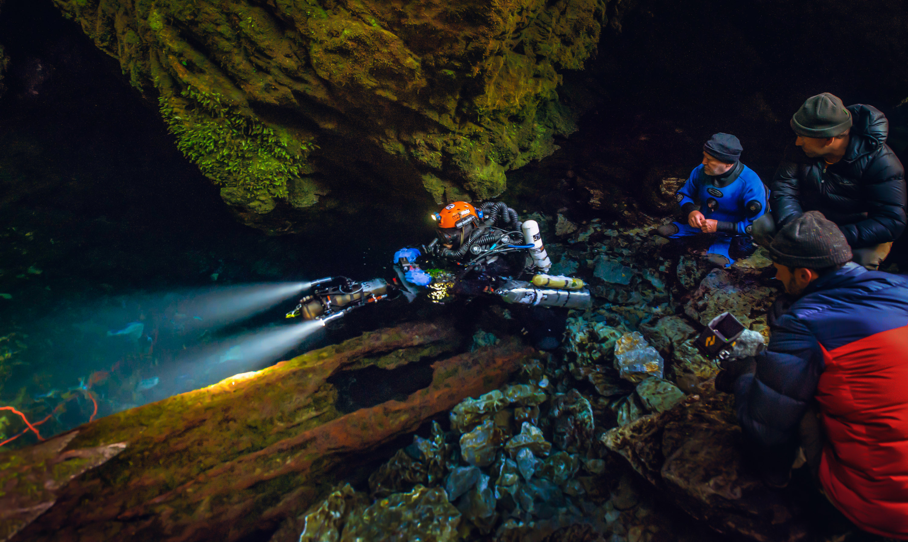 Harris at the mouth of a cave in diving gear with 3 men to the right looking on.